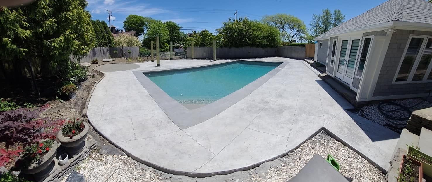 A backyard pool with gray concrete surrounding it, with trees and a house in the background on a sunny day.
