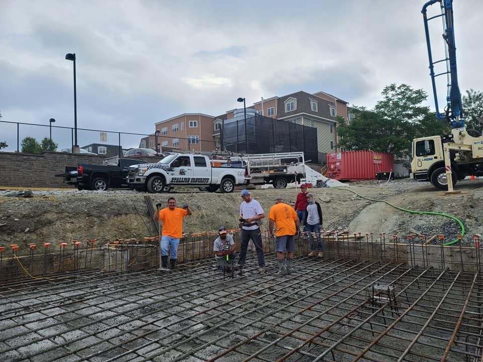 Construction workers on a concrete foundation, preparing to pour. Trucks, equipment, and residential buildings in the background.