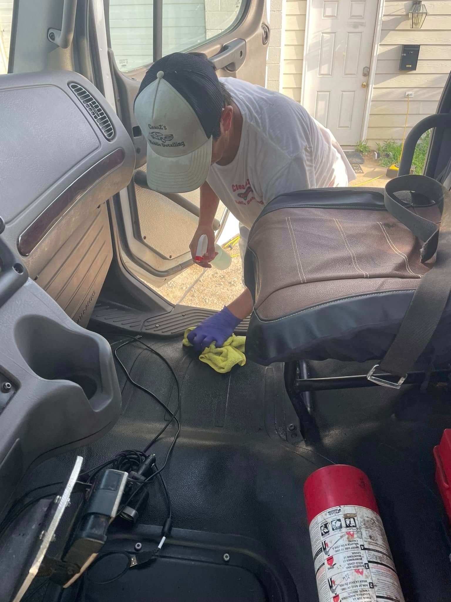 Person cleaning the interior of a vehicle, wearing a hat, gloves, and using a spray bottle and cloth.