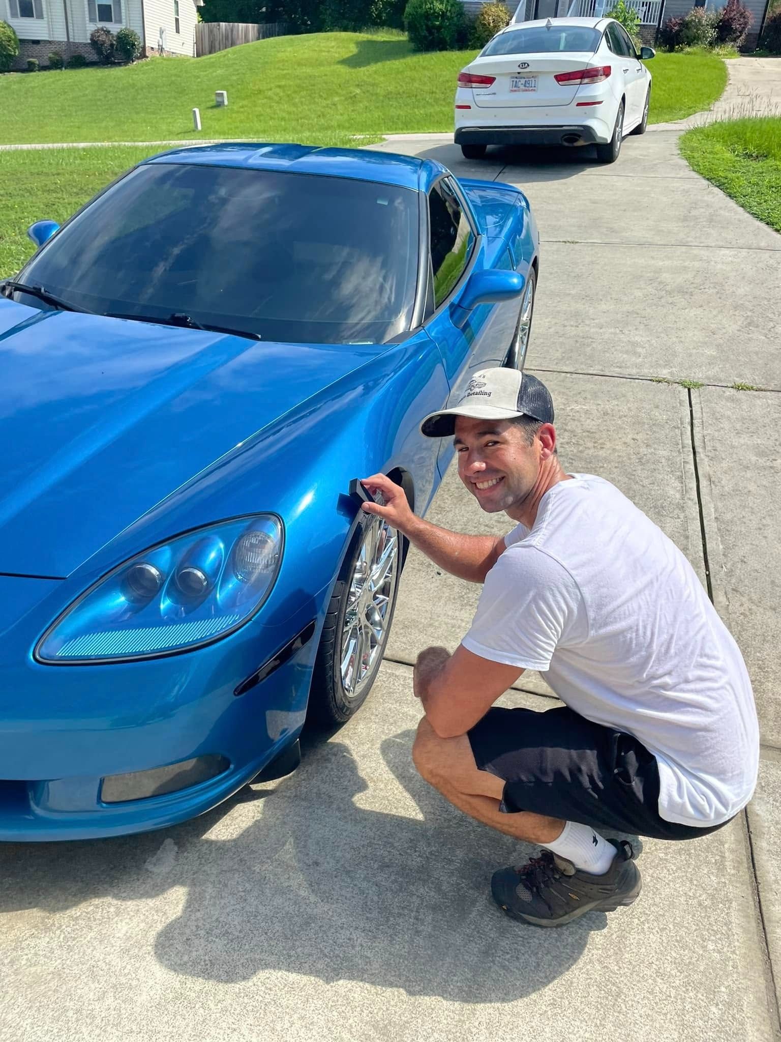 Man crouching next to a blue sports car, smiling. Cars parked on a driveway on a sunny day.