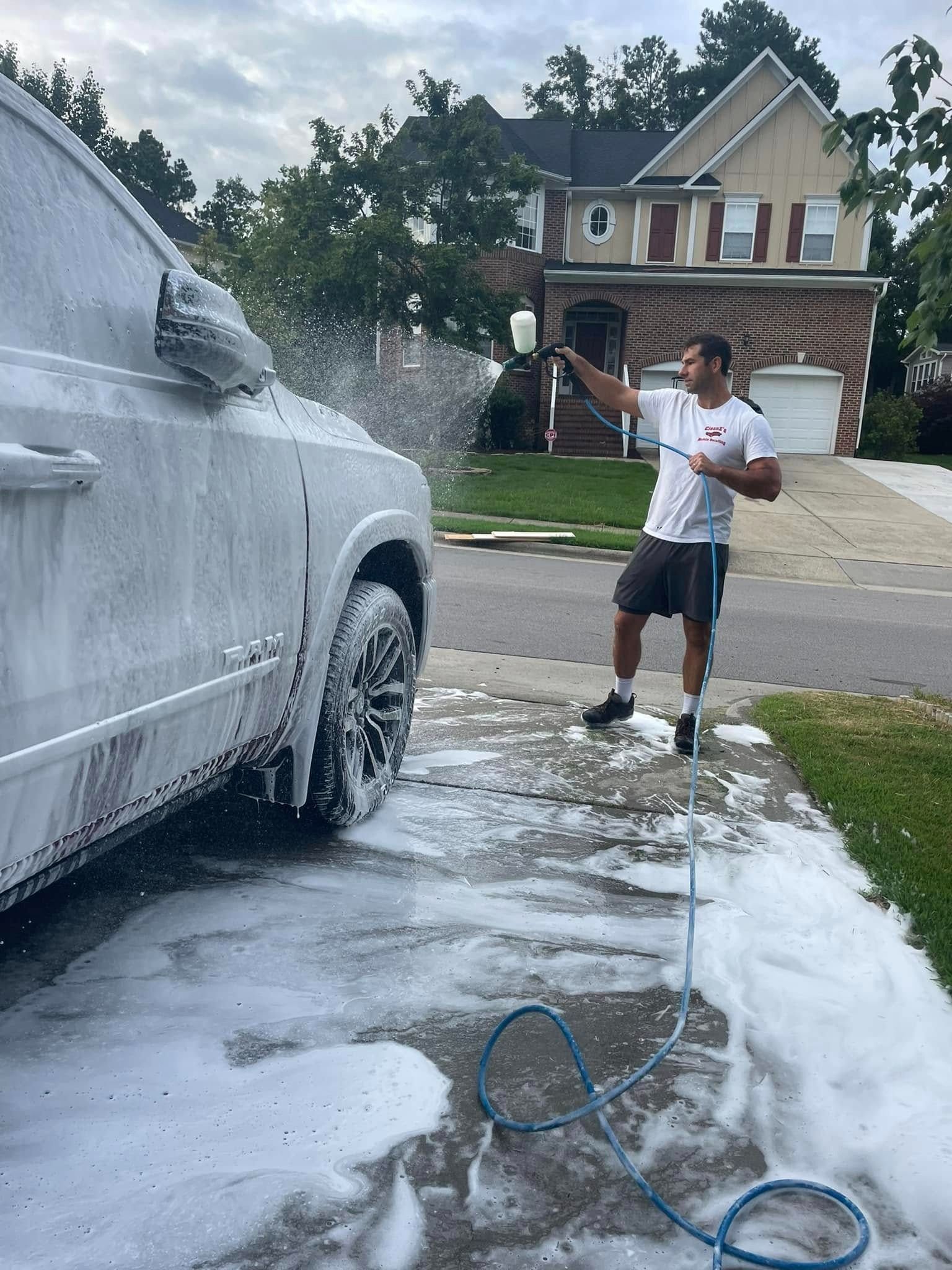 Man washes white SUV with foam in driveway. House in background.