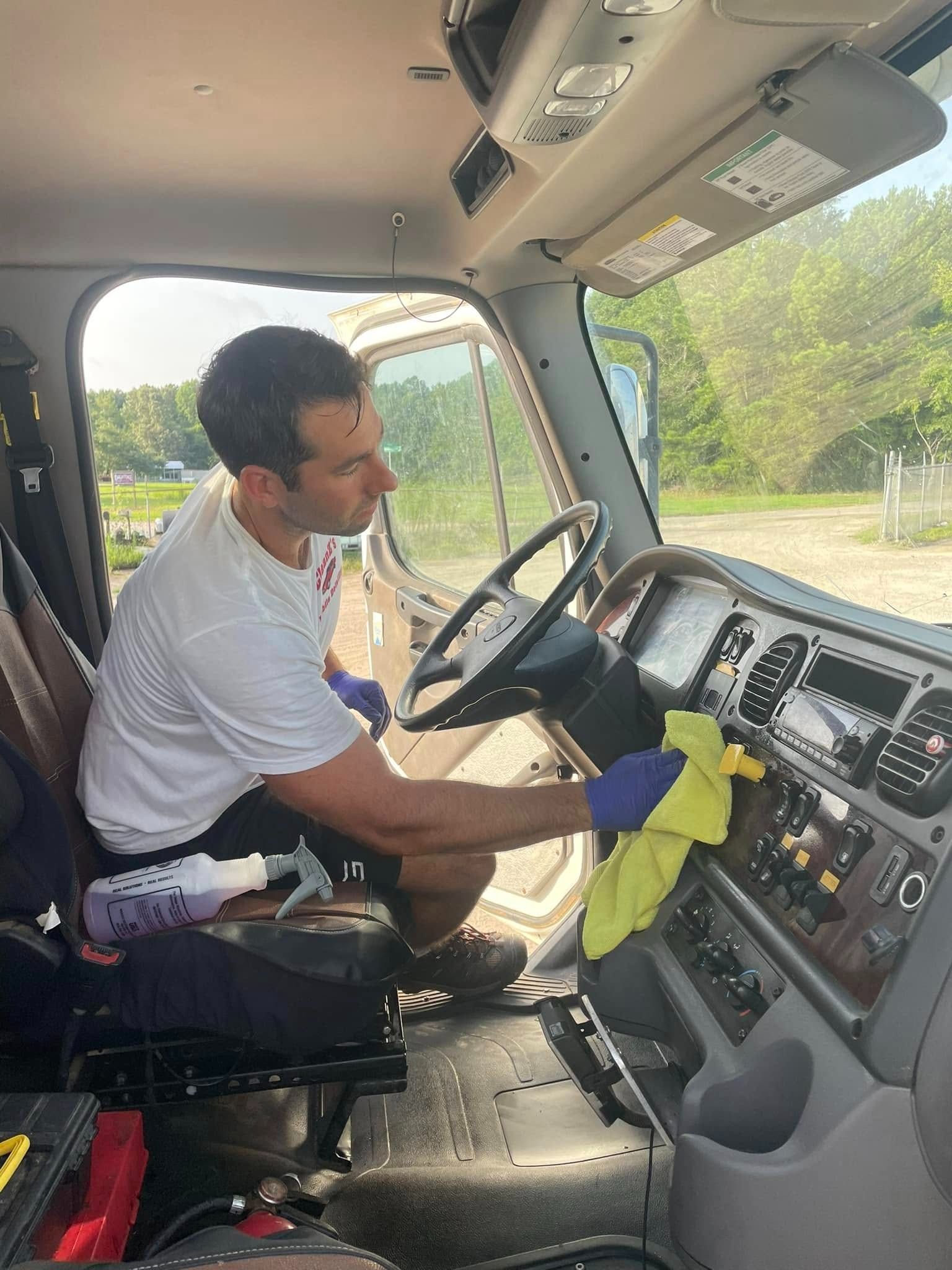 Man in gloves cleaning a truck's dashboard with a yellow cloth. Inside truck, sunny day.
