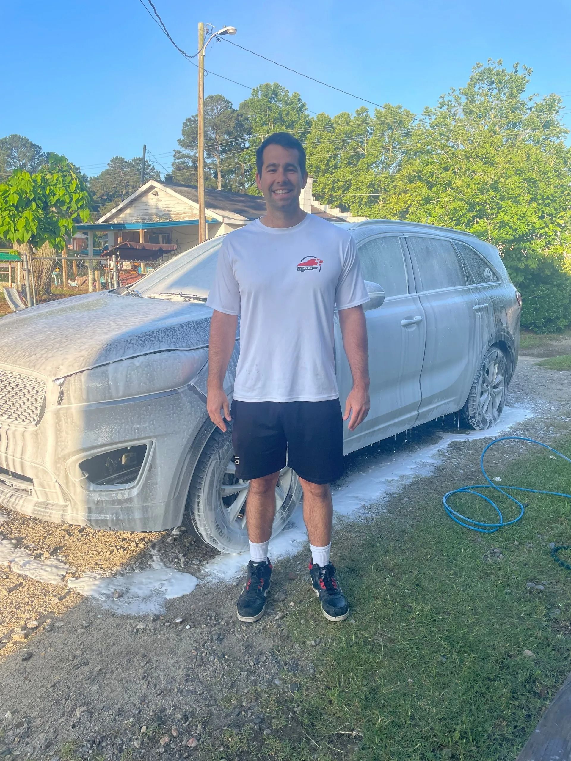 Man washing a gray SUV in a yard. He wears a white shirt, black shorts, and stands smiling.