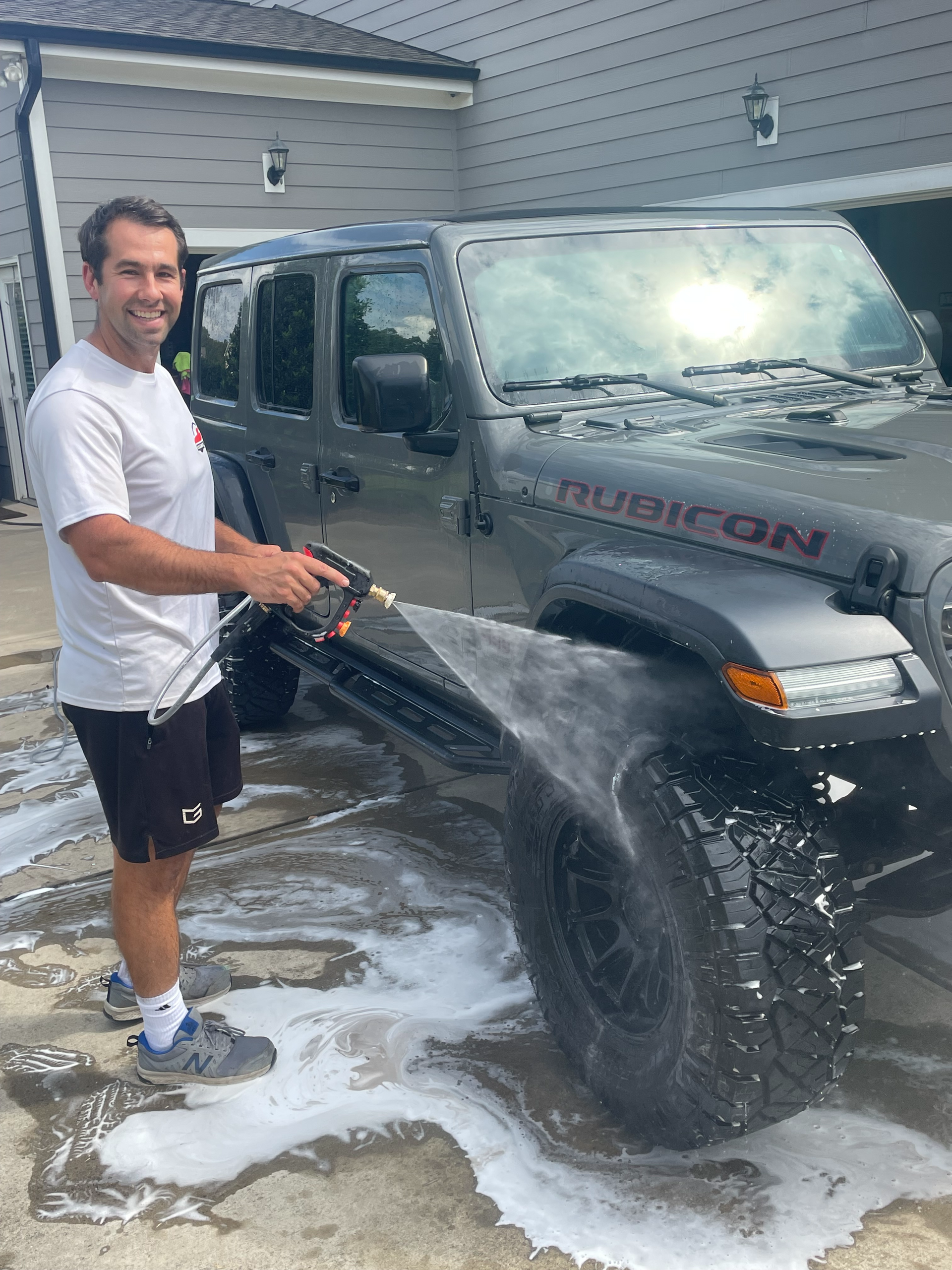 Man washing a gray Jeep Rubicon with soapy water. Outdoors in driveway, smiling.