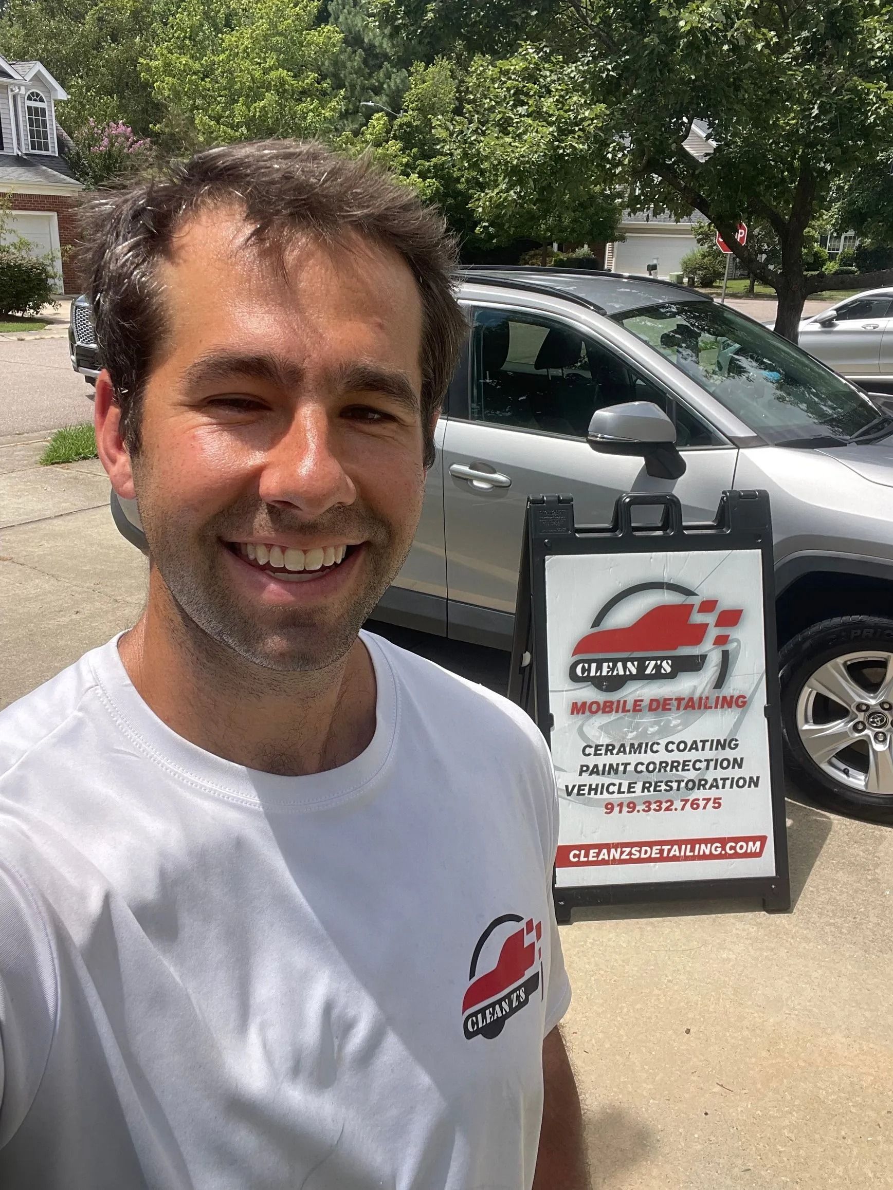 Man smiling, standing near an A-frame sign for car detailing services; silver SUV in the background.
