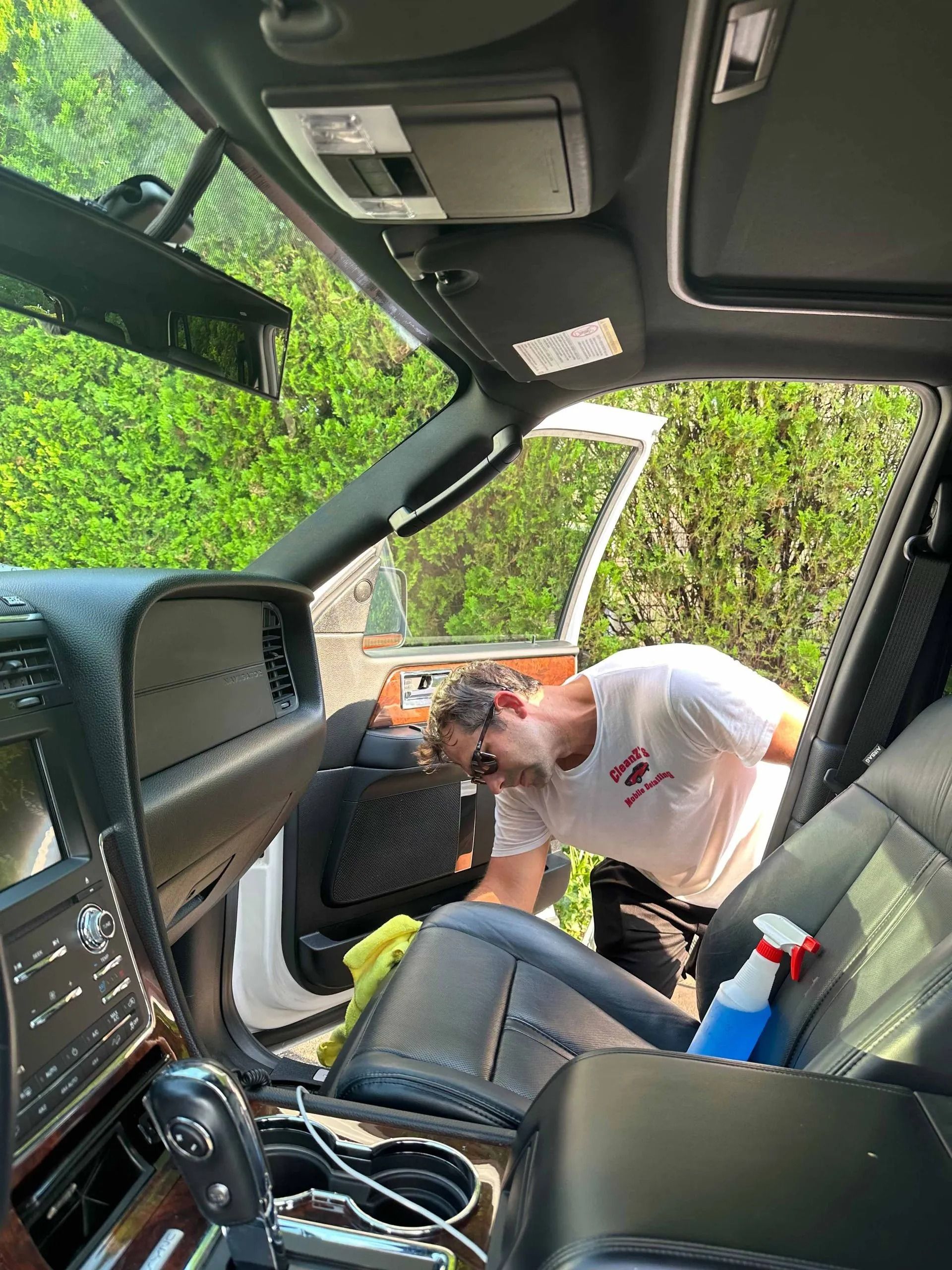 Man cleaning the interior of a white car parked outdoors. He's using a spray bottle and cloth.