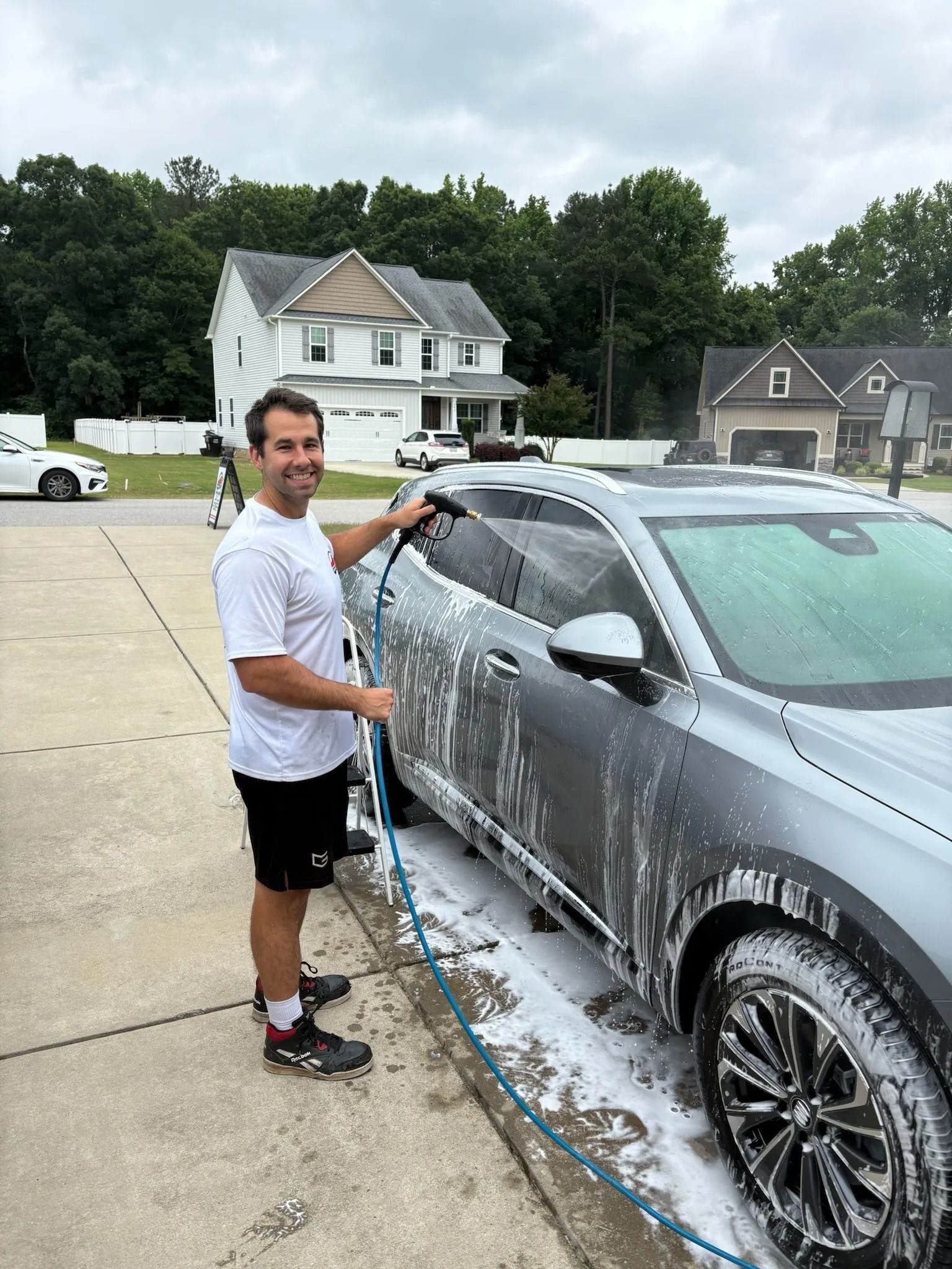 Man washing a gray car in a driveway, smiling, with a house in the background.