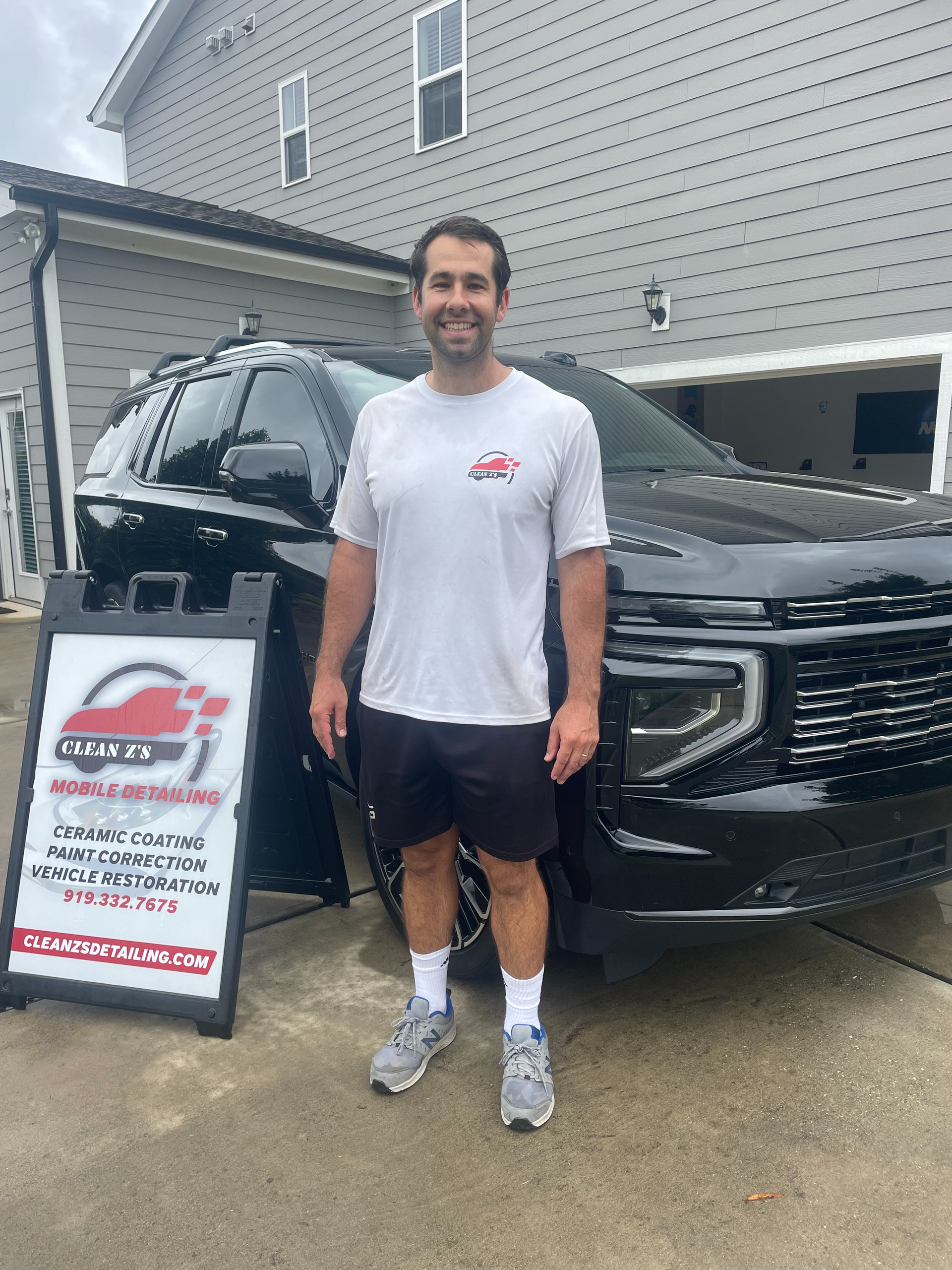 Man stands in front of black SUV, sign for auto services. He wears light shirt, shorts, and sneakers.