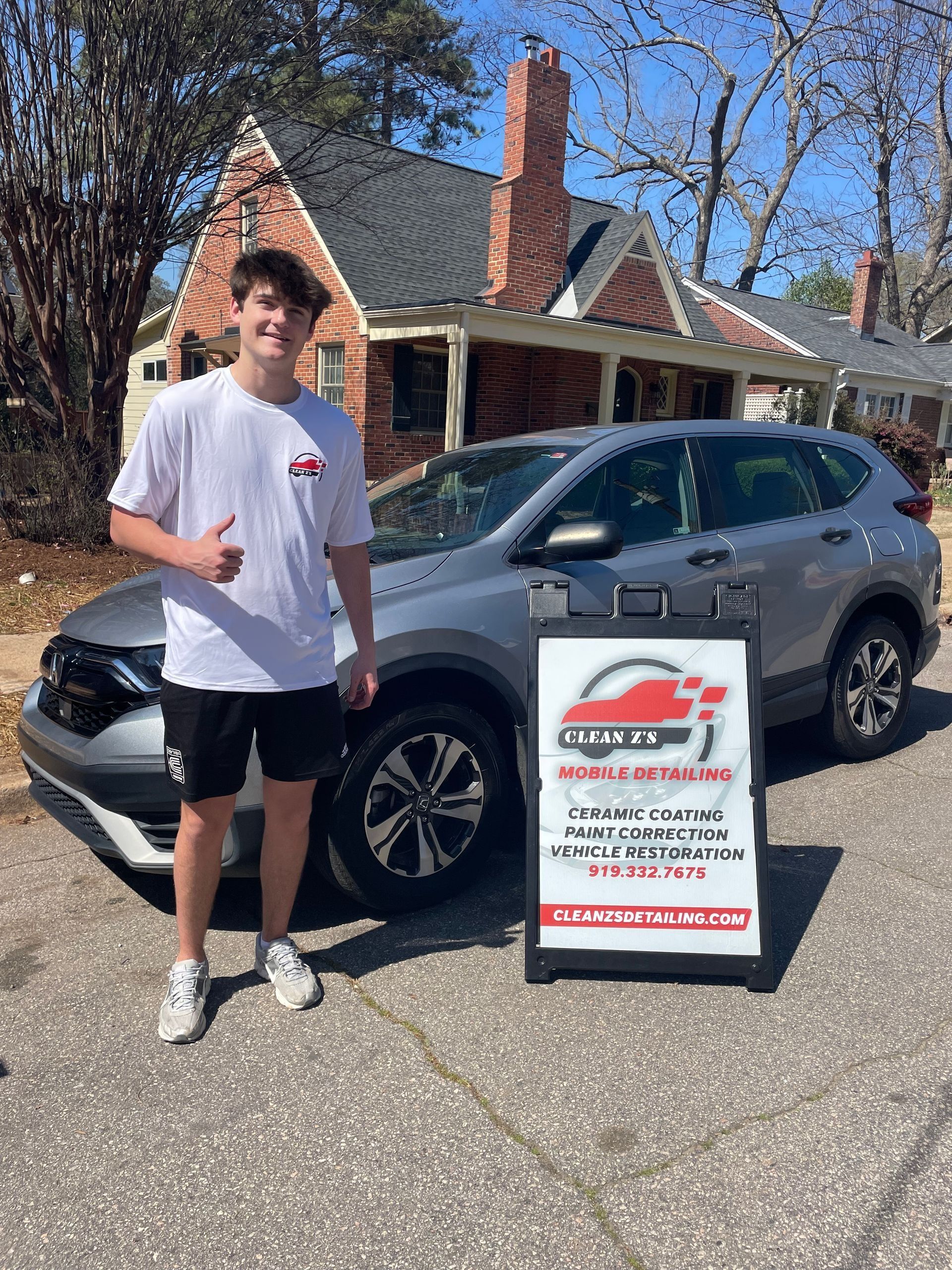 Young man giving a thumbs up next to a gray SUV in front of a house with a sign that has car sales information.