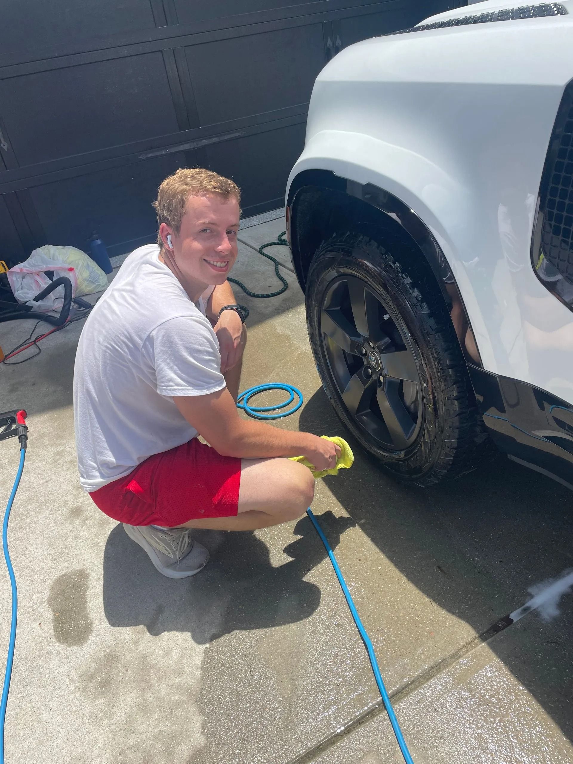 Man in red shorts washing a white car's tire, smiling in the sun.