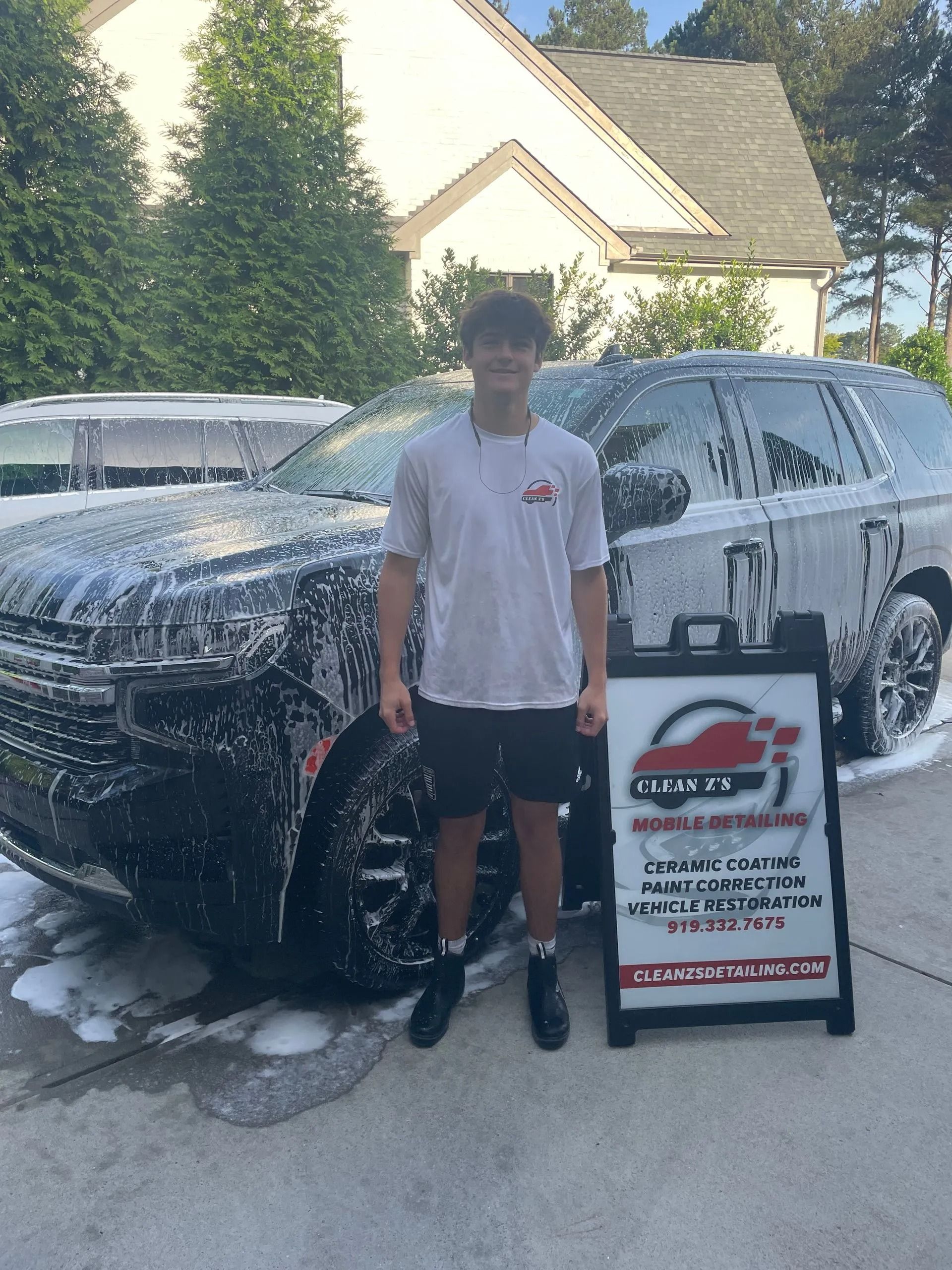 Teenager stands next to a foamy, black SUV at a car wash, holding a sign.