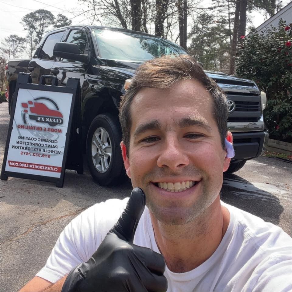 Man in black gloves gives thumbs-up, with a black truck and business sign in the background.