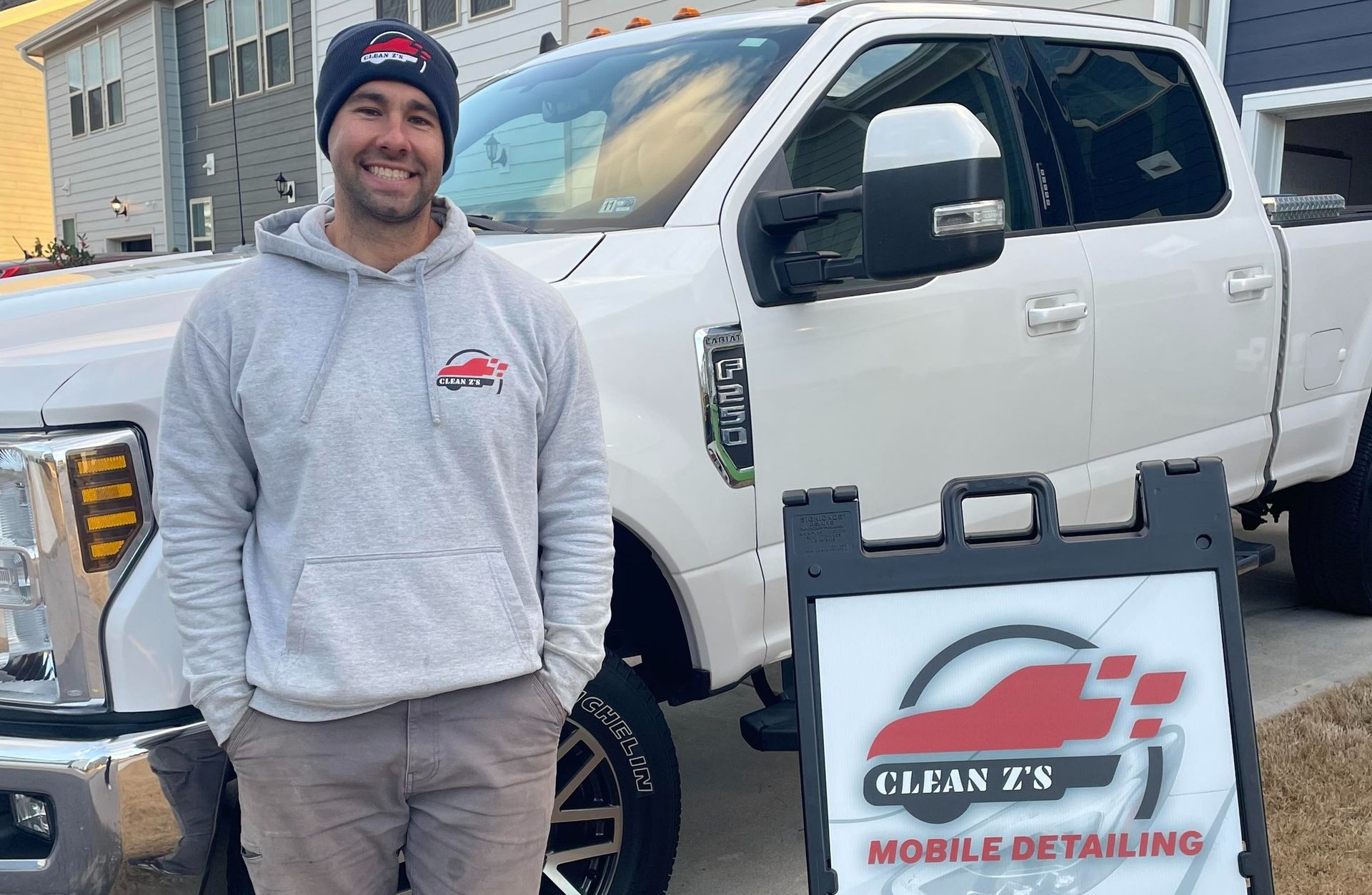 Man in a gray hoodie smiles beside a white pickup truck. Sign reads