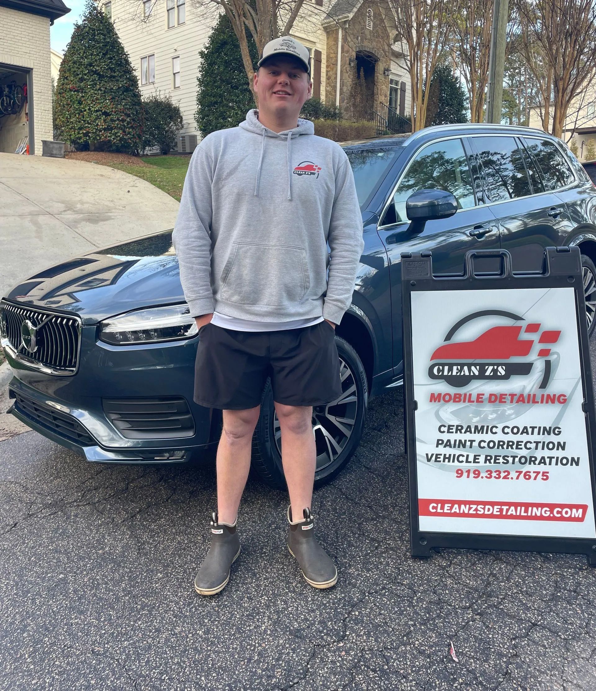 Man in gray hoodie and shorts stands by car, next to a sign for