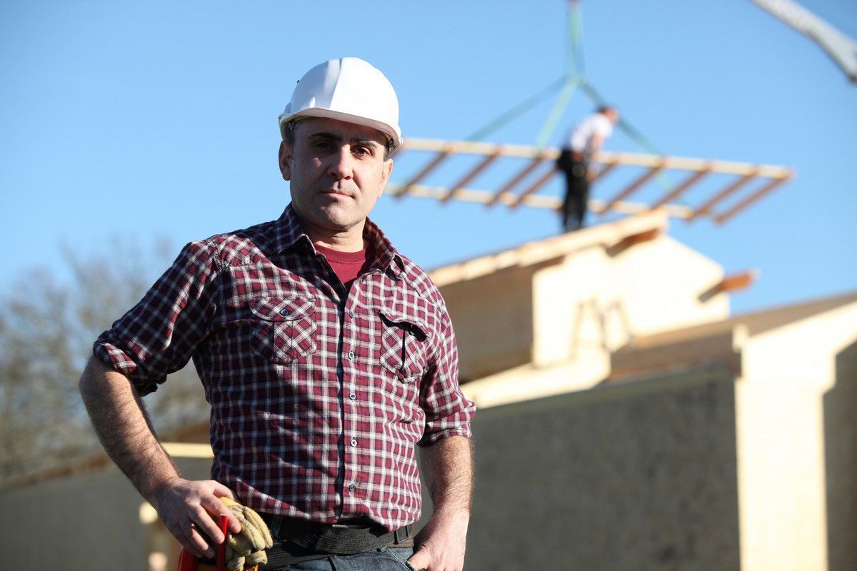 Construction worker in plaid shirt and hard hat, posing in front of building under construction.