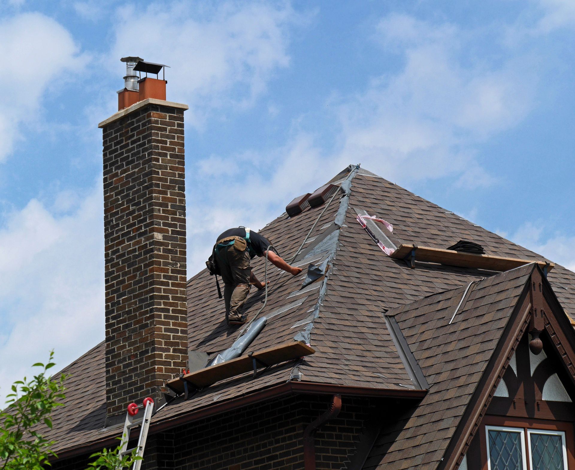 Roofer on a brown shingle roof near a brick chimney, working against a cloudy sky.
