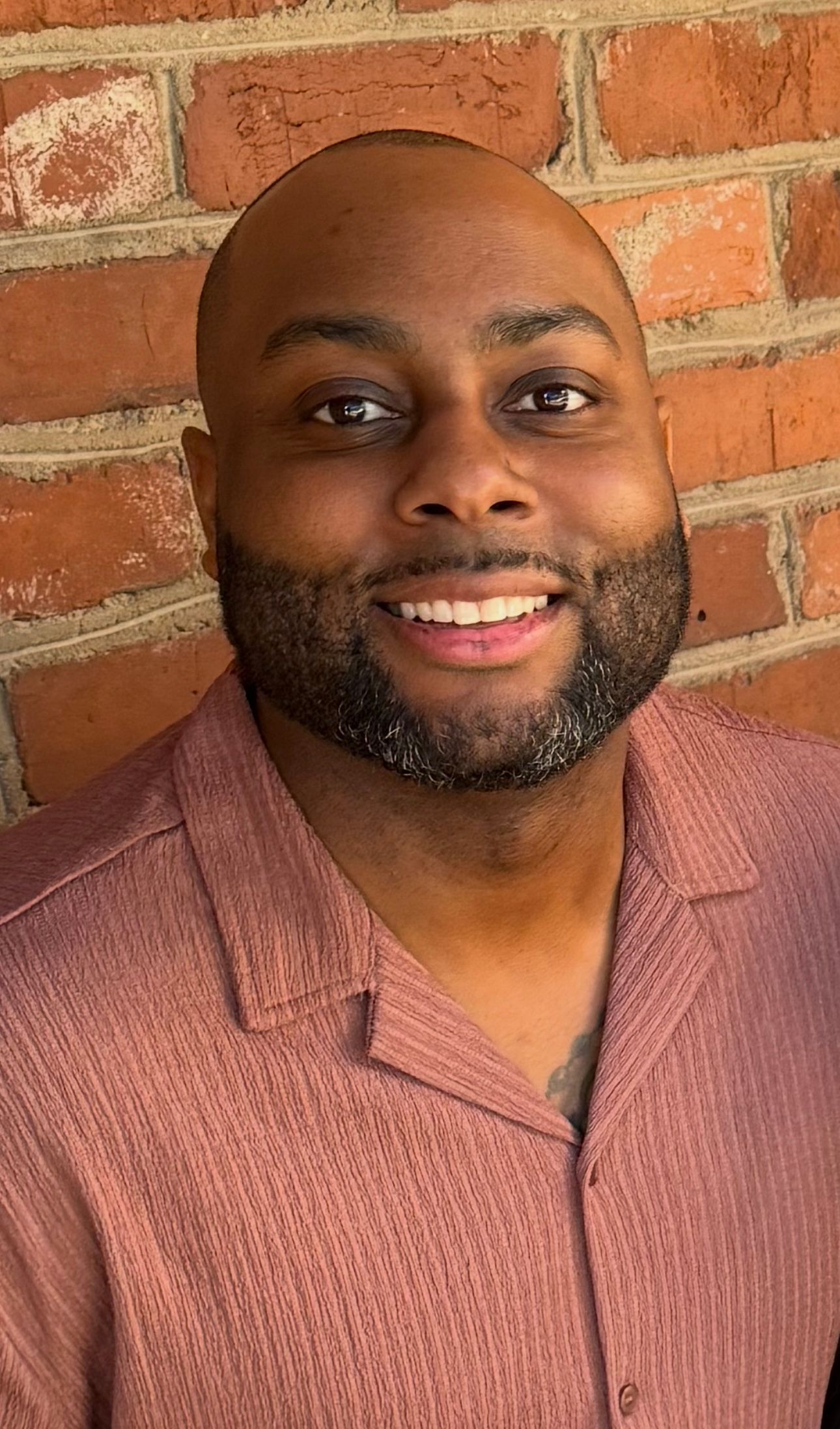 Man with a beard smiling in front of a brick wall, wearing a peach-colored button-down shirt.