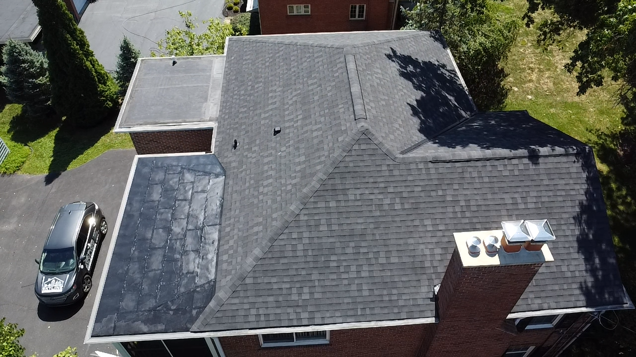 Overhead view of a dark-shingled house roof with a brick chimney, surrounded by trees and a driveway with a car.