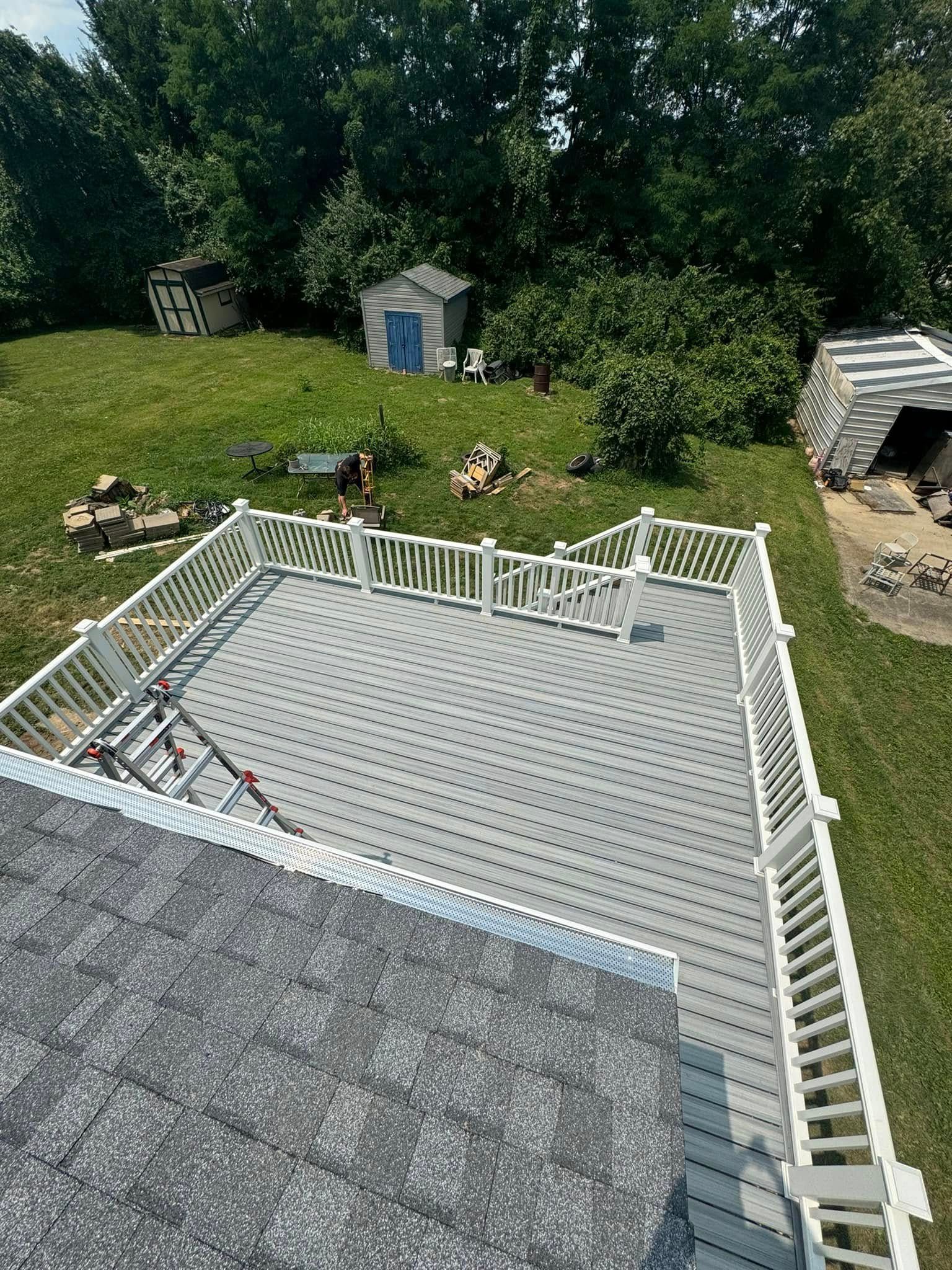 Overhead view of a gray deck with white railing, overlooking a grassy backyard with trees and small sheds.