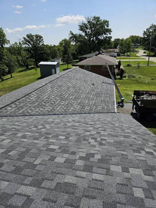 View of a gray shingled roof on a house; background shows trees, other houses, and a blue sky.