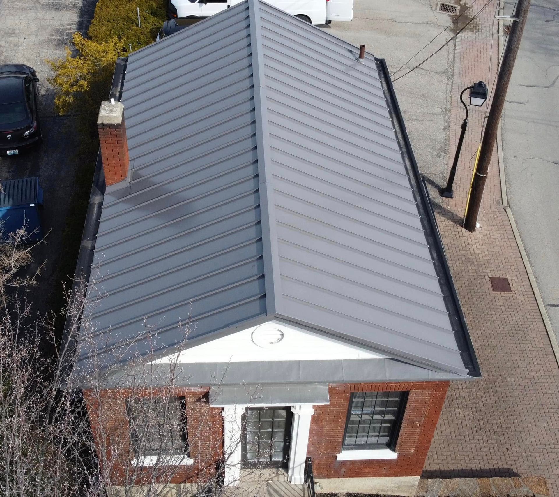 Brick building with grey metal roof, two windows, and chimney. Side view of street with car and tree.