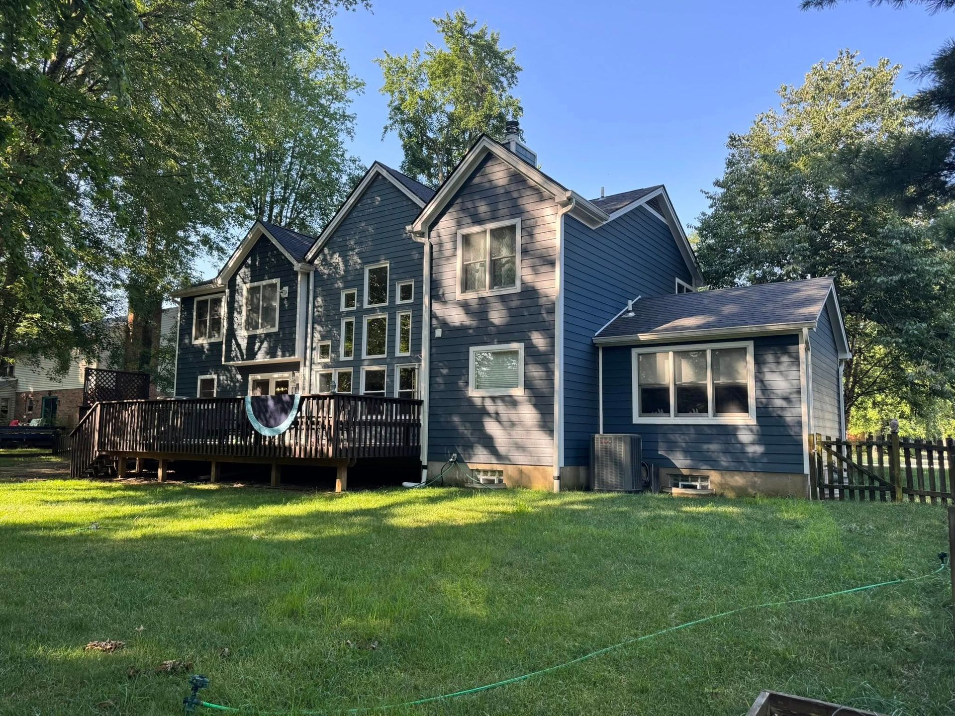 Backyard of a blue house with a wooden deck and green lawn, trees surround.