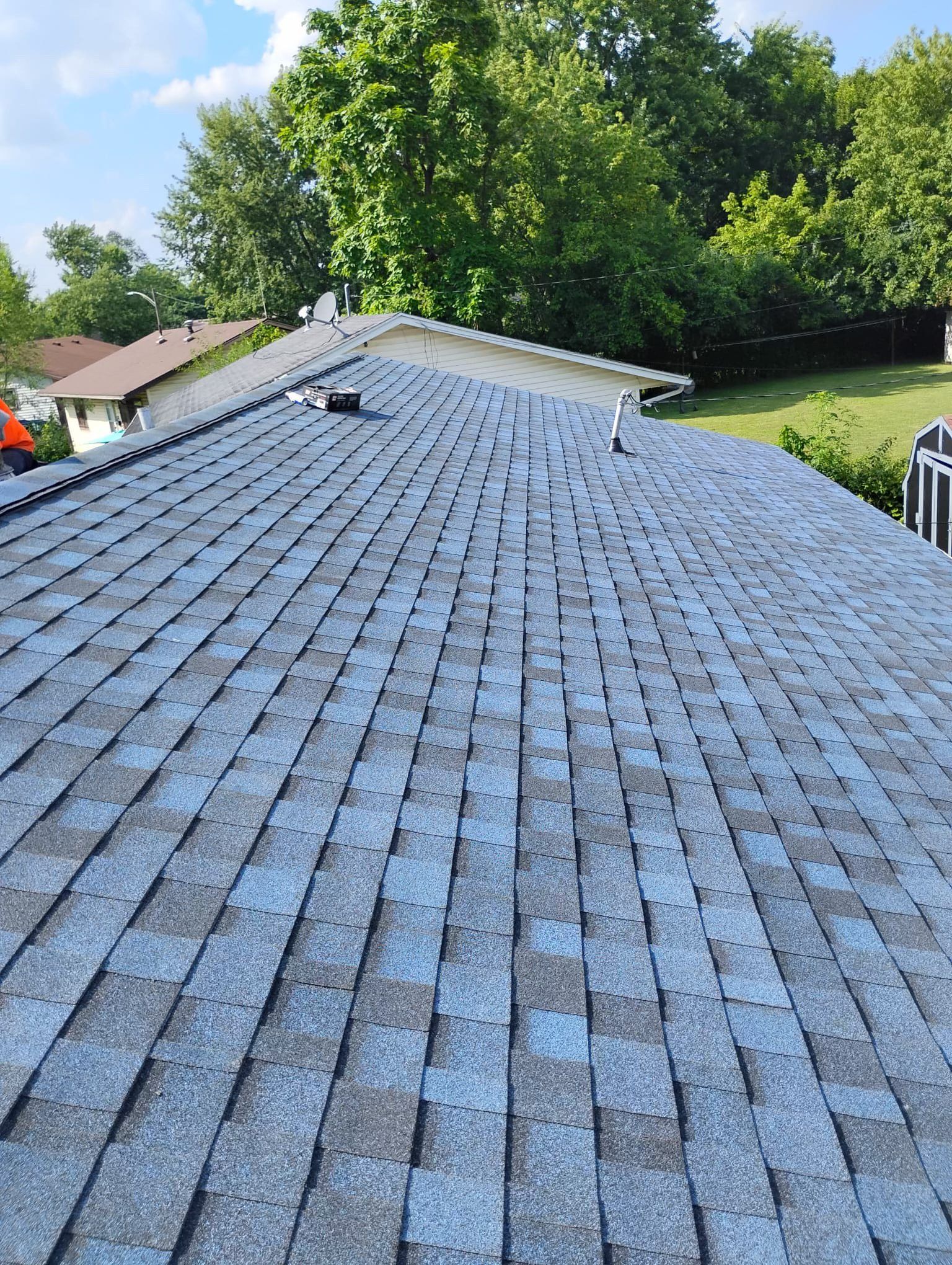 Blue asphalt shingle roof on a house with surrounding trees and blue sky.