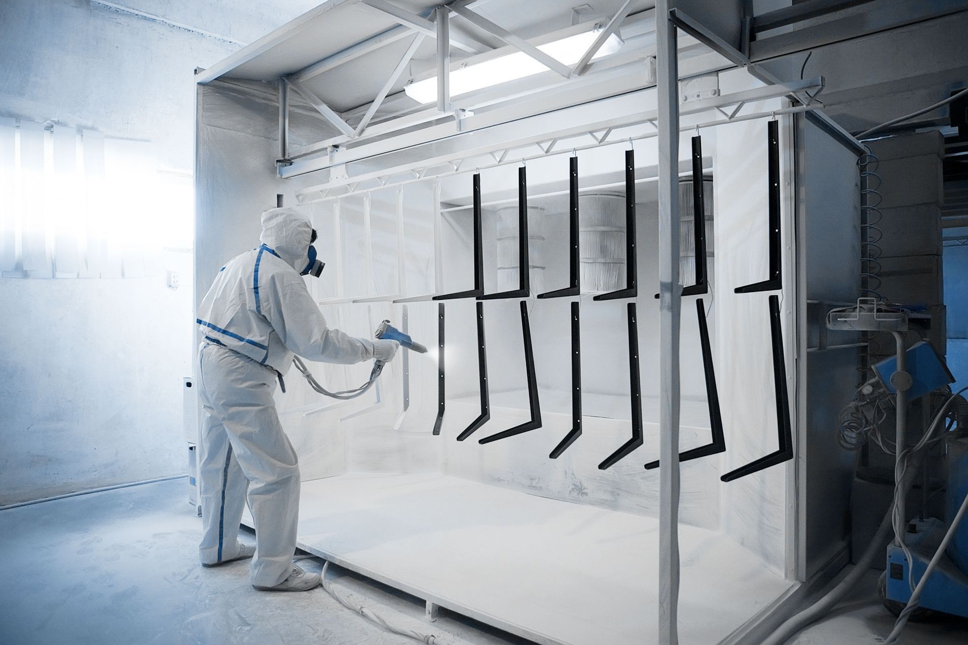 Person in protective suit spray painting black metal brackets in a white spray booth.