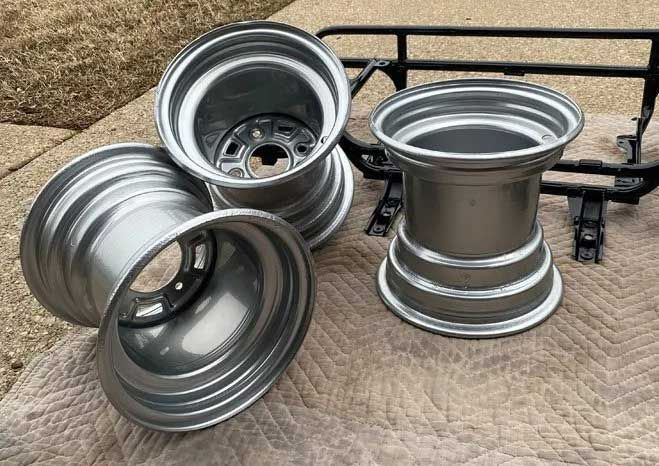 Four silver ATV wheels and a black rack on a patterned surface outdoors.