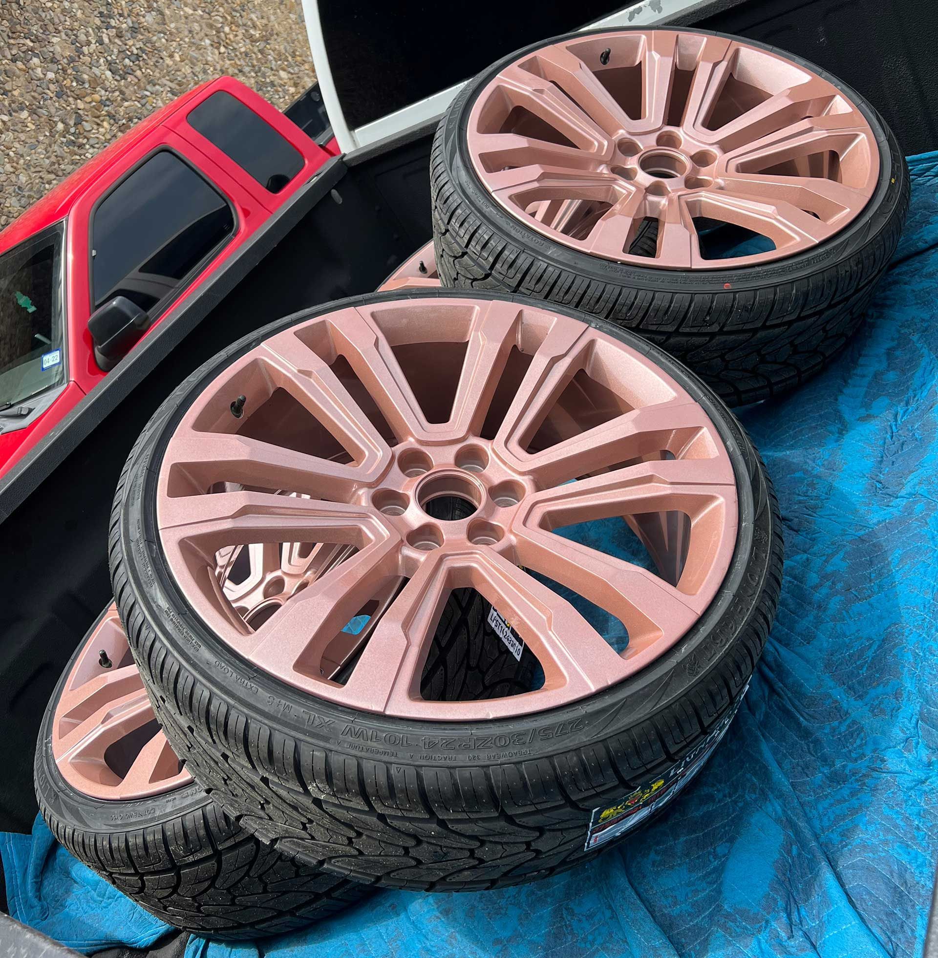 Four car tires with rose gold rims on a blue tarp, next to a red car door.