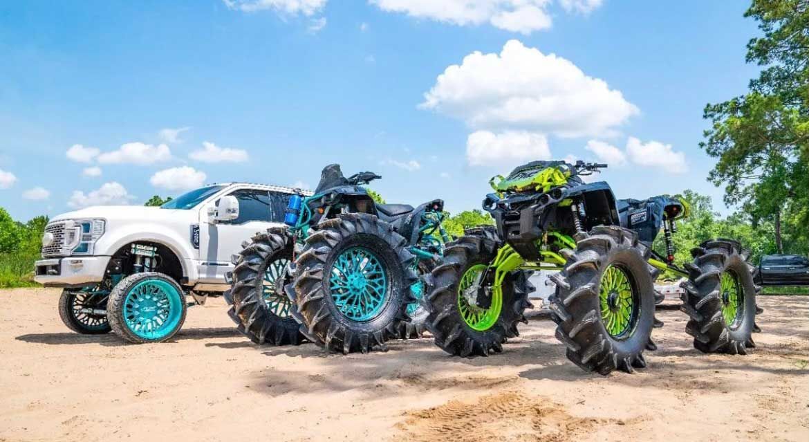 A lifted white truck and two mud-bogging ATVs with large tires on a dirt road under a blue sky.