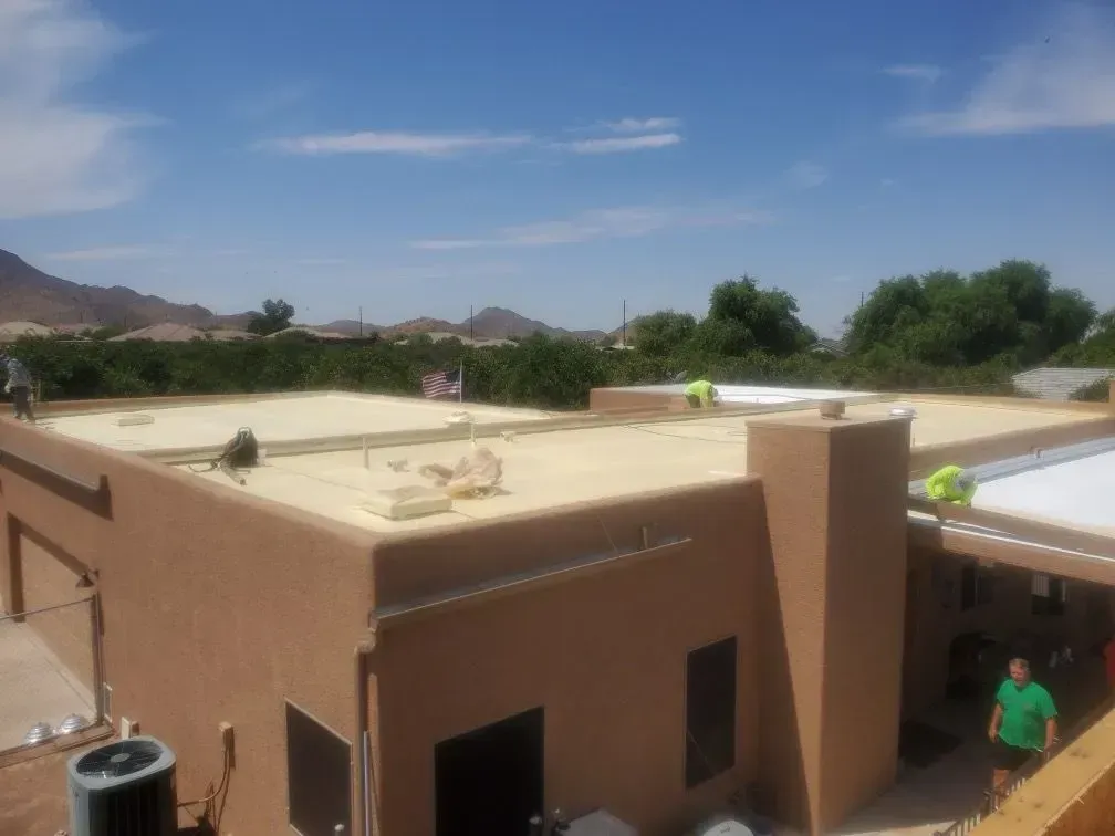 Workers on a flat roof with beige coating; brown building, blue sky, trees in the background.