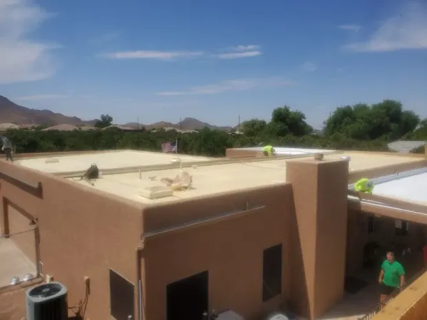 Workers on a flat roof with beige coating; brown building, blue sky, trees in the background.