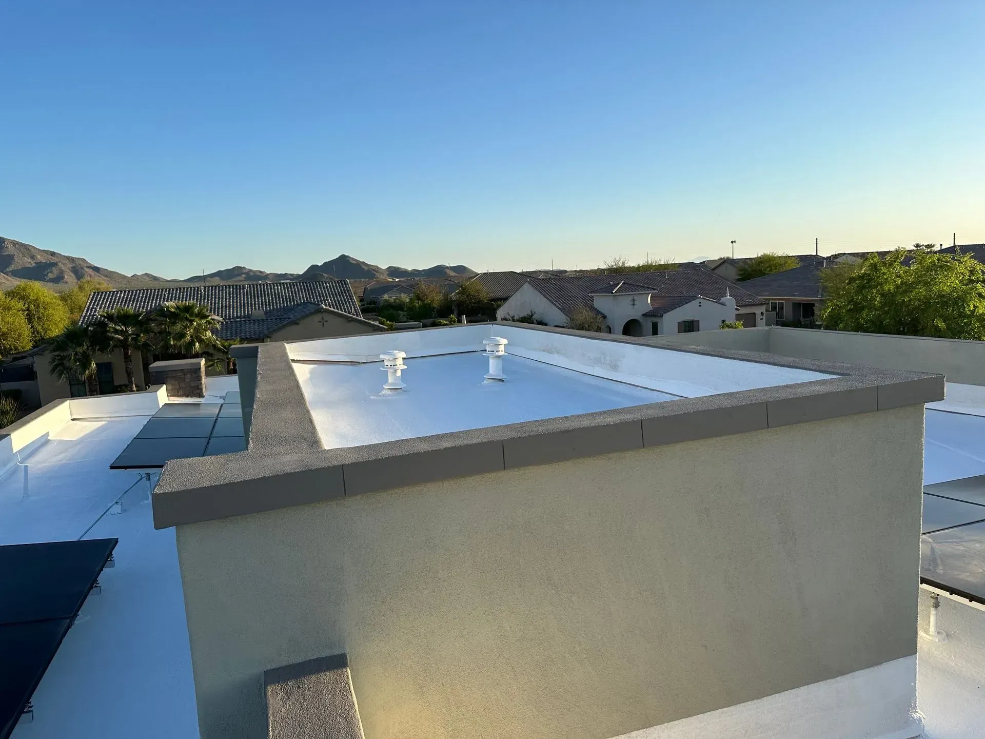View of a flat, white roof with vents, surrounded by rooftops, houses, and mountains under a blue sky.