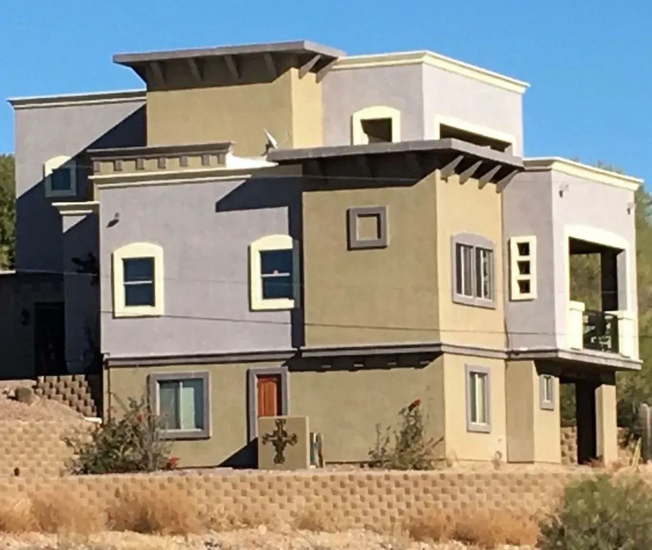 Two-story house with tan, gray, and brown stucco exterior, with square and rectangular window openings, and a small front yard with retaining wall.