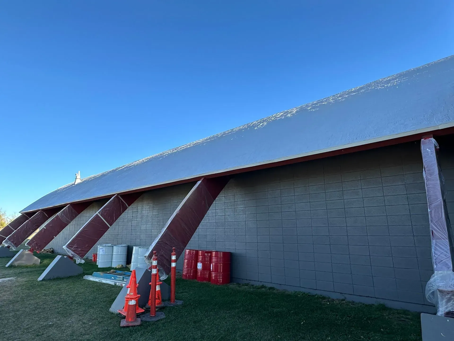 Gray building with angled red supports. Barrels and equipment on the ground. Clear blue sky.