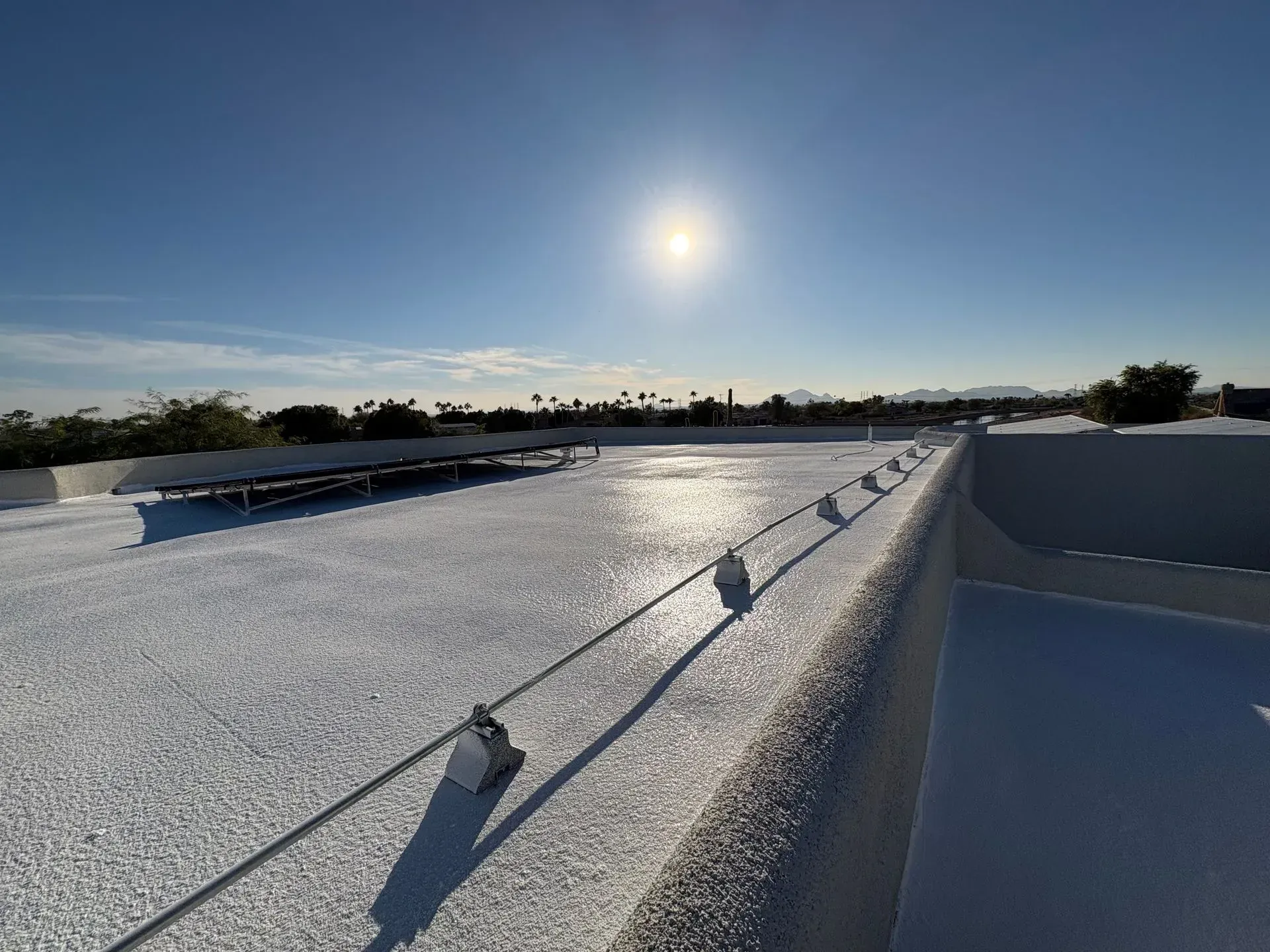 Bright sun over a white rooftop with a cable railing. Distant trees and mountains under a blue sky.