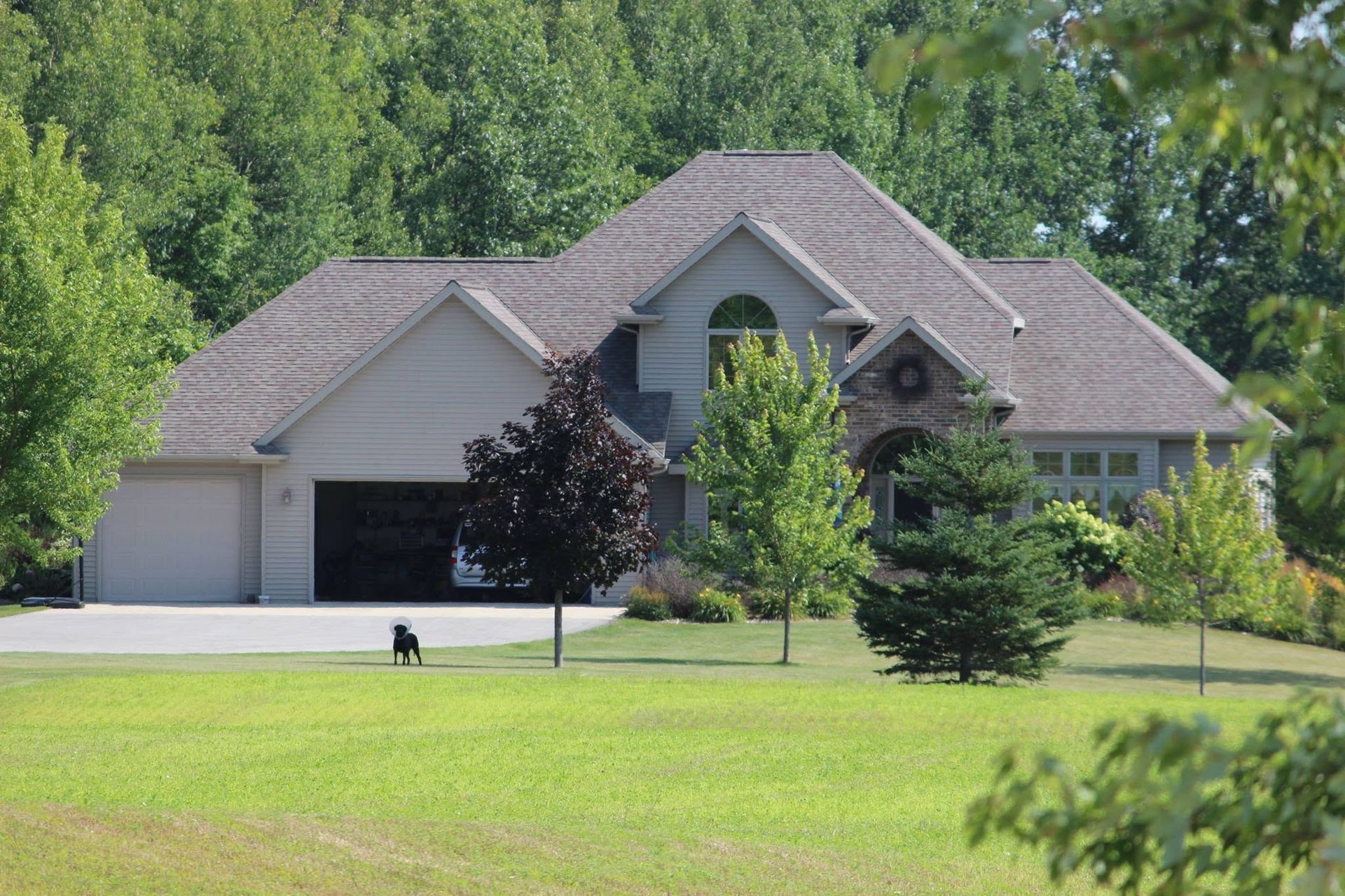 Large gray suburban house with a gabled roof, driveway, and green lawn bordered by trees.