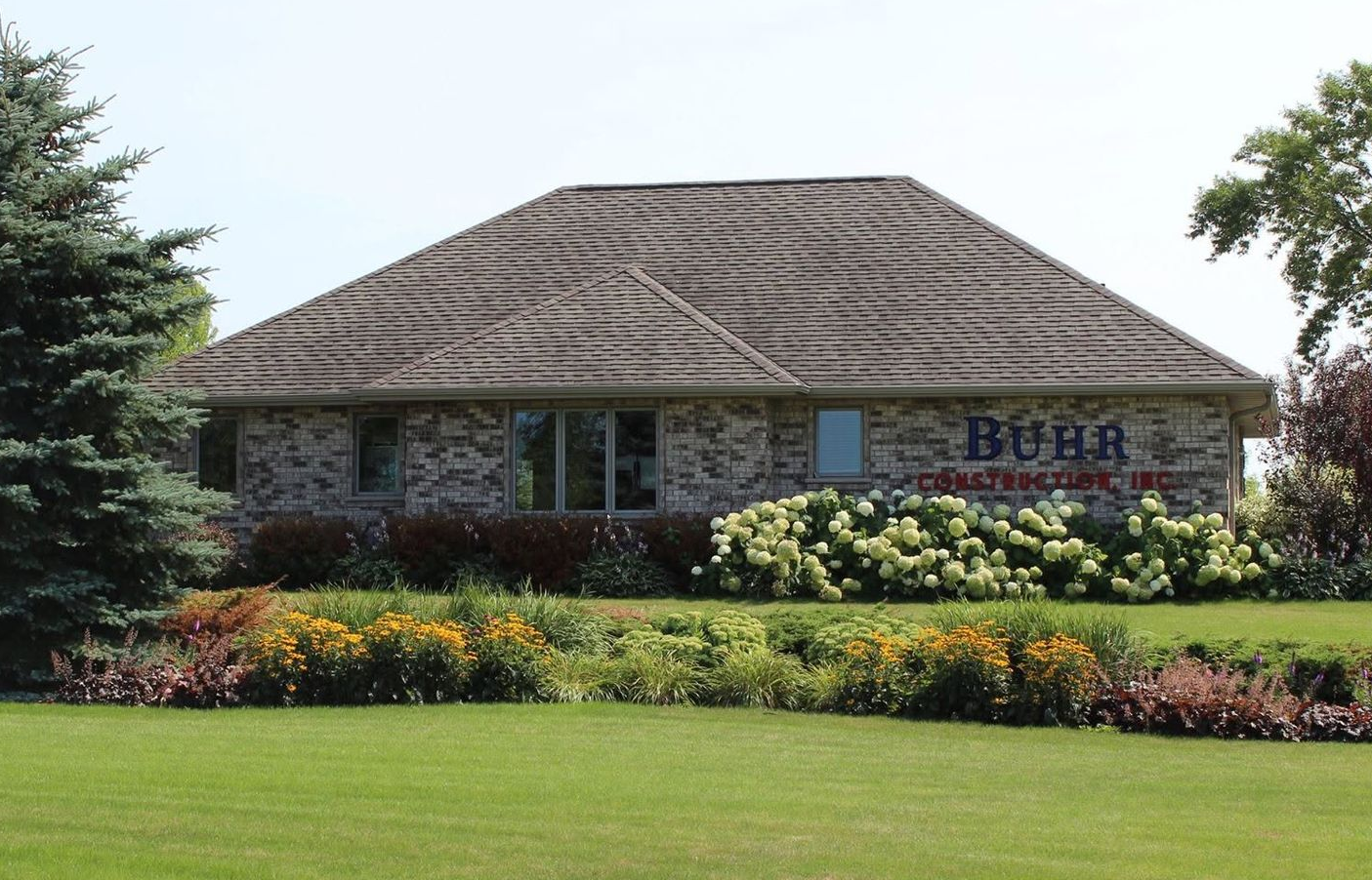Brick building with a striped roof, flowering shrubs, and a grassy lawn under a clear sky