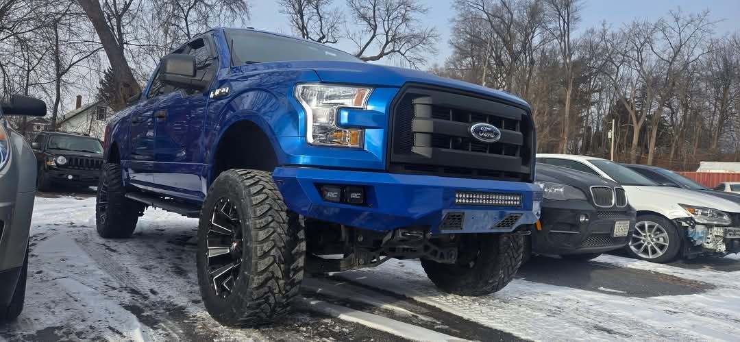 Blue Ford F-150 truck with large tires on snow-covered ground, parked outside, other cars visible.