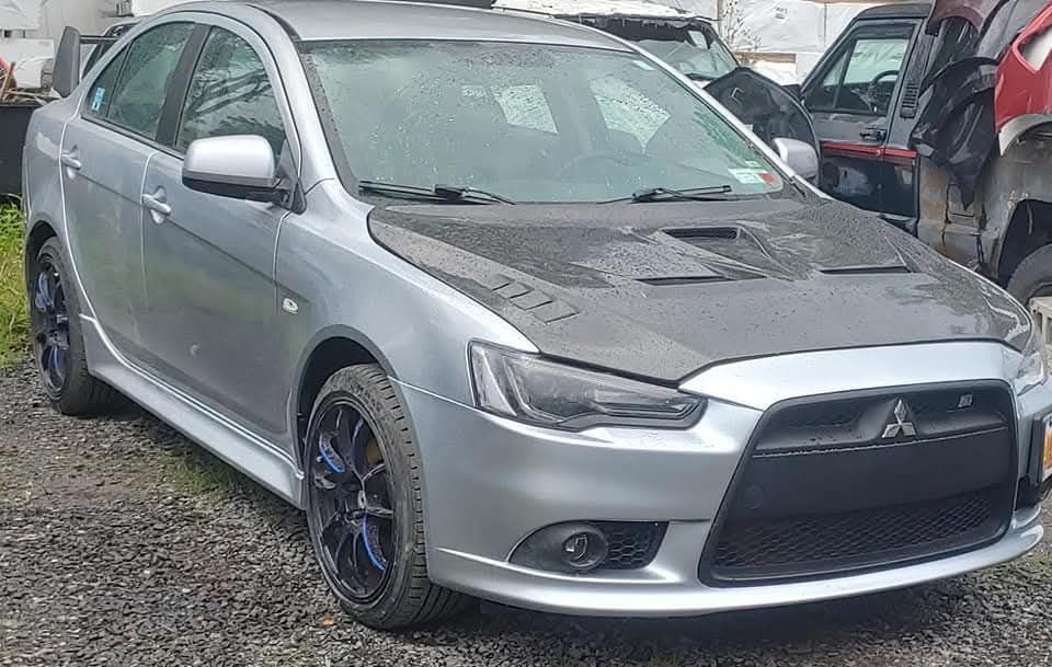 Silver Mitsubishi Lancer with black hood, blue wheels, parked outdoors.