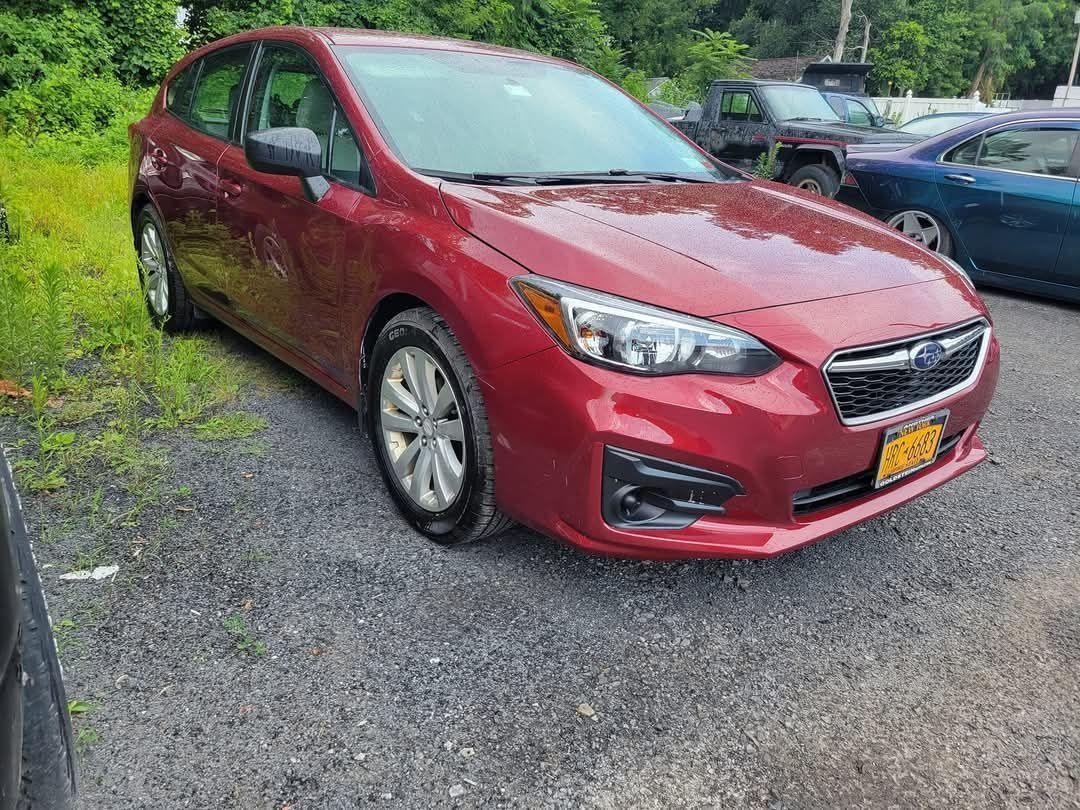 Red Subaru Impreza hatchback parked on gravel, surrounded by grass and other vehicles.