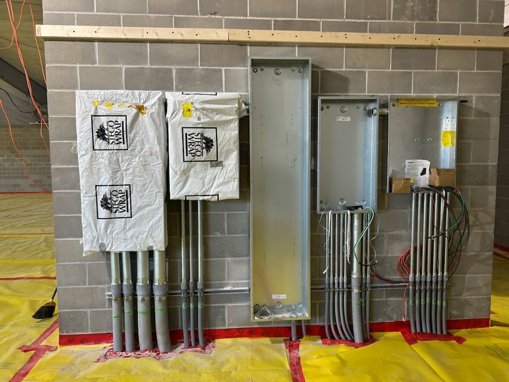 Electrical panels and conduit mounted on a block wall, some covered in white protective wrapping.