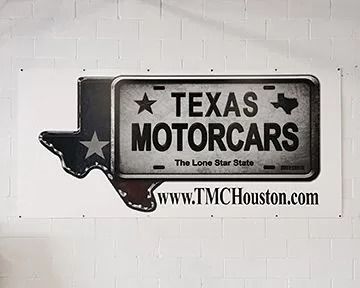 Sign for Texas Motorcars with Texas outline and license plate design. Website address www.TMCHouston.com.