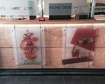 Two Campari ads on a wooden bar: one with red abstract art, the other with a bottle pouring into a glass.