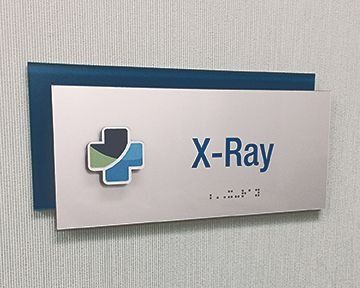 X-Ray room sign with logo, blue and green medical cross, on silver rectangular panel, mounted on blue backing, on textured wall.