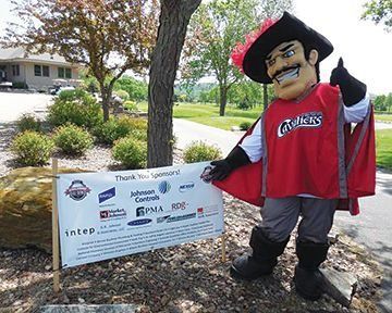 Mascot in red cape and hat with banner thanking sponsors at outdoor event.