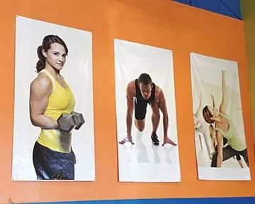 Three fitness posters on an orange wall: woman with dumbbells, man in starting position, and person stretching.