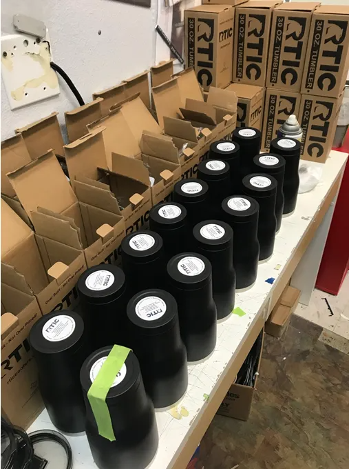 Black tumblers with white labels line a table, cardboard boxes in the background.