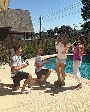 Two men kneel, proposing to two women by a pool. All appear surprised and happy outdoors.